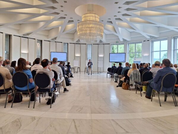 Conferentie in een moderne zaal met veel mensen en een spreker vooraan.
